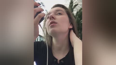 Media: Video of a young woman with fair skin and light brown hair, wearing a black top, using a smartphone while in an indoor setting with green plants in the background.