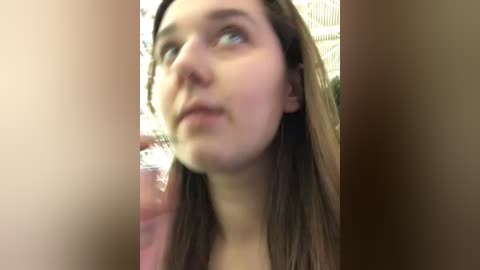 Media: A video of a young woman with fair skin and straight brown hair, wearing a pink shirt, looking upward with a contemplative expression. The background is blurred, featuring a warm, indoor setting.