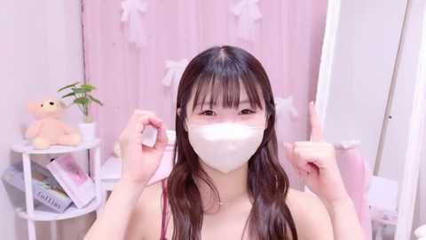 Media: A video of an East Asian woman with long, dark hair and fair skin, wearing a white face mask and a pink top, posing with raised hands in a pastel-pink, softly lit room with decorative elements and plush toys.