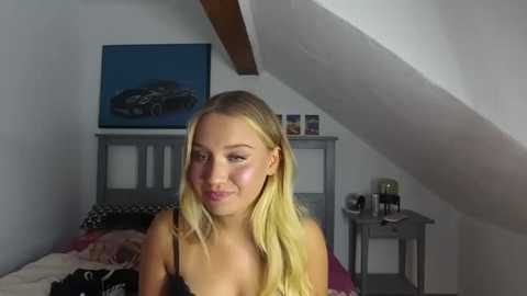 Media: A video of a young blonde woman with light skin and shoulder-length hair, smiling, wearing black lingerie, in a dimly lit attic bedroom with a gray bed, a painting of a boat, and a nightstand.