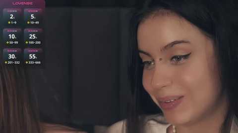 Media: Video of a close-up of a young woman with long black hair, fair skin, and a slight smile, wearing a white top, in a dimly lit room.