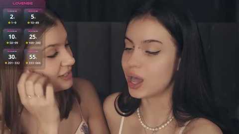 Media: Video of two young women, one with straight, dark hair and a pearl necklace, the other with straight, light brown hair, both wearing light-colored tops, smiling intimately, set in a dimly lit room.