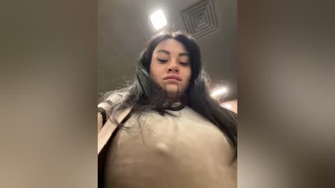 Media: Video of a young woman with long black hair, wearing a beige coat, taken from a low angle, showing her chest and face. The background features a ceiling with a circular light fixture.