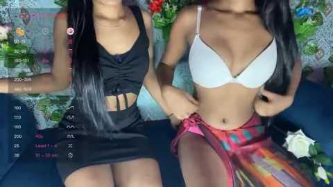 Media: Video of two women with medium brown skin, wearing black and white lingerie, posing seductively on a blue couch. Background features a floral patterned wall.