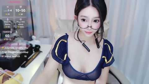 Media: A video of a young East Asian woman with long black hair, wearing a sheer blue bra with yellow accents, sitting at a desk with a computer screen displaying a live stream.