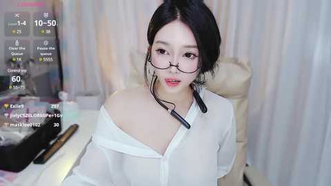 Media: Video of a young Asian woman with fair skin, black hair, and glasses, wearing a white off-shoulder blouse, sitting at a desk with a microphone, against a soft-focus background.