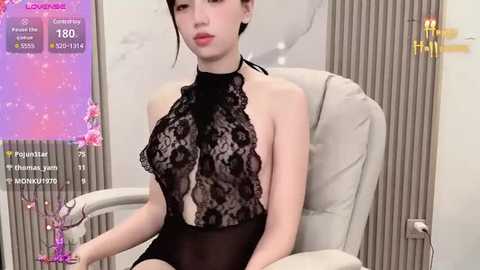 Media: A video of a young Asian woman with fair skin, wearing a revealing black lace bodysuit, sitting in a white chair. The background features a pink, starry digital overlay and beige curtains.