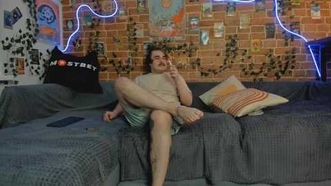 Media: Video of a man with a mustache, wearing a tank top and shorts, sitting on a gray couch in a cozy room with a brick wall adorned with posters and neon lights.