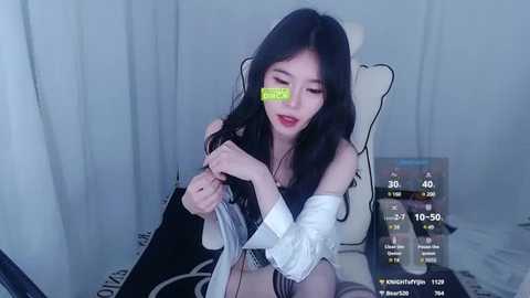 Media: Video of an East Asian woman with long black hair, wearing a white off-shoulder dress, sitting in a gaming chair, covered by a green sticker on her face.