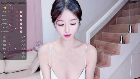 Media: Video of an Asian woman with fair skin, dark hair in a bun, wearing a white camisole, standing in a modern, light-toned living room with a beige sofa and a wooden staircase.