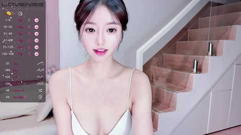 Media: Video of an Asian woman with pale skin, dark hair styled in a bun, wearing a white satin bralette, standing indoors beside a staircase.