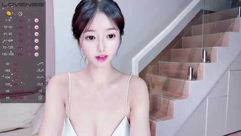 Media: A video of an East Asian woman with pale skin and long, dark hair in a bun, wearing a white, low-cut camisole. She stands indoors next to a staircase with wooden steps and a glass railing.