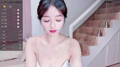 Media: Video of a young Asian woman with fair skin, dark hair in a bun, wearing a white camisole, standing near a wooden staircase in a modern, minimalist home.