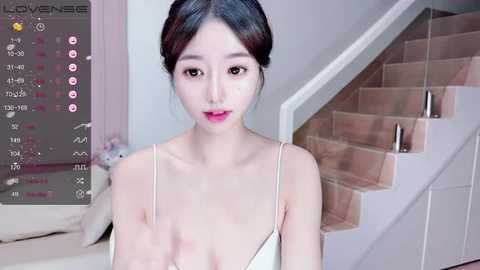 Media: Video of a young East Asian woman with fair skin, dark hair in a bun, wearing a white spaghetti-strap top, standing on a beige staircase in a modern, well-lit room.