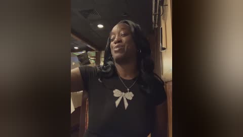 Media: A video of a smiling African-American woman with long black hair, wearing a black t-shirt with white ribbon appliqu\u00e9s and a silver necklace. She stands in a dimly lit, cozy room with wooden walls and ceiling lights.