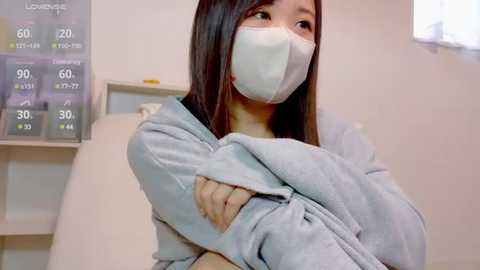 Media: Video of an Asian woman with long black hair, wearing a white face mask and gray hoodie, hugging herself, in a sterile, light-colored medical room with a temperature gauge on the wall.