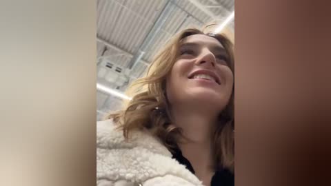 Media: Video of a smiling young woman with blonde hair, wearing a fur coat, in a warehouse with exposed pipes and white walls.