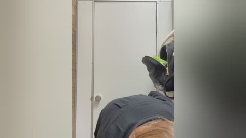 Media: Video of a person wearing a black jacket and a green helmet, looking through a frosted glass door with a silver handle, in a dimly lit hallway with beige walls.