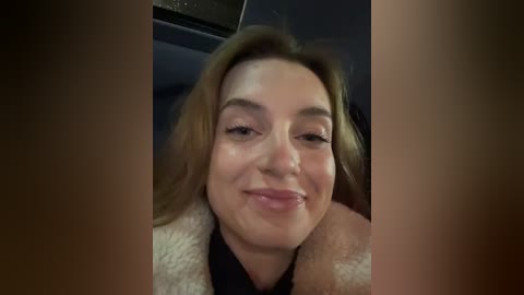 Media: Video of a smiling, light-skinned woman with light brown hair, wearing a fur-lined jacket, seated in a dimly lit car.