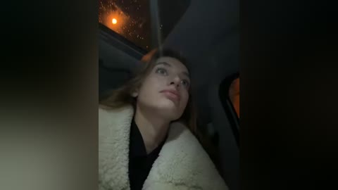 Media: A video of a young Caucasian woman with light skin and shoulder-length brown hair, wearing a fluffy white cardigan, gazing out the car window at a lit streetlamp, creating a soft, ambient glow.