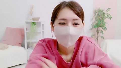 Media: Video of an East Asian woman with brown hair, wearing a pink hospital gown and a surgical mask, sitting on a white couch in a bright, minimalistic room with light-colored walls and a plant.