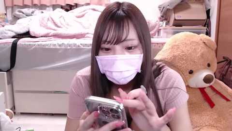 Media: Video of an Asian woman with long brown hair, wearing a face mask, pink top, and pink nails, playing with a smartphone in a cozy, cluttered bedroom with a teddy bear.