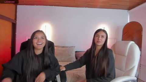 Media: Video of two identical twins, both with long dark hair, wearing black jackets, seated on white chairs in a warmly lit room with a wooden ceiling and soft furnishings.