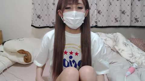 Media: Video of an Asian woman with long brown hair, wearing a white mask, shirt, and shorts, sitting on a bed with a pink vibrator beside her.