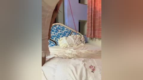 Media: Video of a messy bed with white sheets, a blue and white patterned pillow, and a pink curtain in the background, creating a chaotic and unmade bedroom scene.