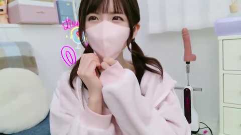 Media: A video of a young East Asian woman in a pink hoodie, wearing a pink face mask, holding a pink heart emoji, in a room with a white bed and a pink vibrator.