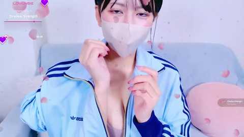 Media: A video of an Asian woman with pale skin, wearing a light blue jacket, unzipped to reveal a bra, and a white face mask, against a pastel background with heart stickers.