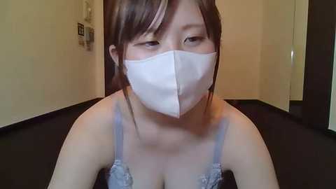 Media: A video of an Asian woman with fair skin and brown hair, wearing a white surgical mask, blue lace bra, and a white tank top, in a dimly lit room with beige walls and a black floor.