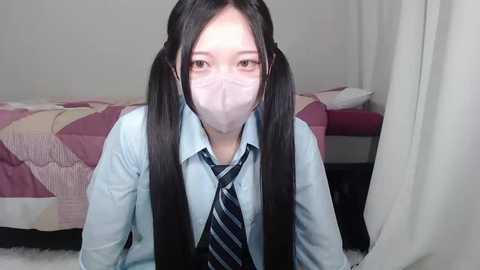 Media: Video of an Asian woman with long black pigtails, wearing a light blue button-up shirt, a dark blue striped tie, and a white face mask, kneeling on a white carpeted floor in a bedroom with a bed and a pink quilt.