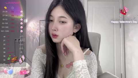 Media: Video of an Asian woman with long black hair, wearing a white lace top, looking pensive, in a dimly lit room with a Valentine's Day theme.