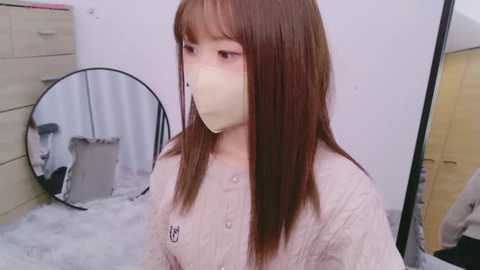 Media: Video of a young Asian woman with straight brown hair, wearing a beige, button-up shirt, and a beige face mask, standing in a cluttered bedroom with a round mirror and beige dresser.