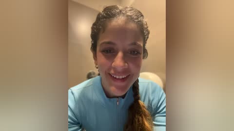 Media: Video of a smiling woman with medium-brown skin and dark hair styled in a braid, wearing a light blue athletic top, standing in a beige-walled hallway.