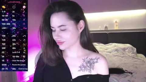 Media: Video of a young woman with long black hair, fair skin, and a detailed floral tattoo on her shoulder, smiling softly in a bedroom with a messy bed and purple lighting.