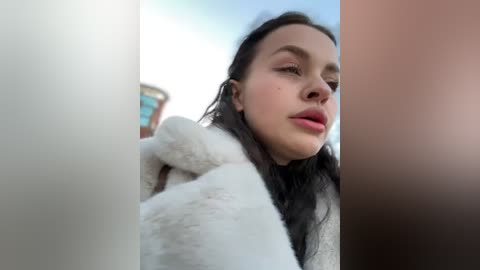 Media: Video of a young woman with long, dark hair and fair skin, wearing a white fur coat, looking introspective against a blurred urban background.