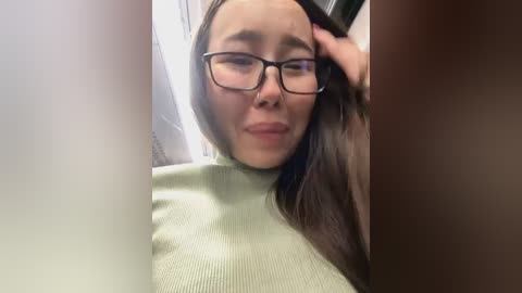 Media: Video of a young woman with light skin, straight brown hair, and glasses, wearing a light green sweater, looking tired and holding her head. Background is blurry, possibly an indoor setting.