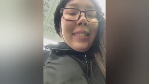 Media: Video of a smiling Asian woman with glasses, wearing a black jacket, in a dimly lit room, possibly an office or indoor setting.