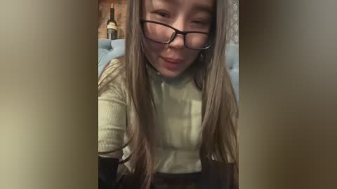 Media: Video of a smiling, light-skinned Asian woman with long, straight, blonde hair, wearing black-rimmed glasses and a cream-colored turtleneck sweater. She is indoors, with a brick wall and a wine bottle in the background.