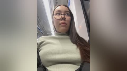 Media: Video of a young woman with long, straight brown hair and glasses, wearing a ribbed, light green turtleneck sweater, sitting in a modern, clean subway train with metal bars and blue lighting.