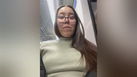 Media: Video of a young woman with long, straight brown hair, wearing glasses and a light green sweater, standing inside an airplane cabin. The image is slightly blurred and out of focus.