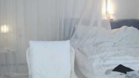 Media: A video of a serene, dimly-lit bedroom with white sheer curtains, a fluffy white comforter, and a white pillow. The background features a white wall and soft lighting, creating a tranquil, minimalist ambiance.