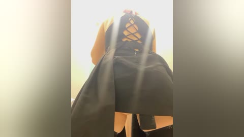 Media: Video of a person in a yellow and black costume with a laced back, bending over. The background is blurred, with a warm, soft lighting effect.