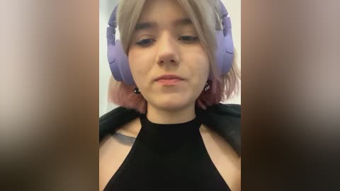 Media: A video of a young woman with light skin and short, pastel pink hair, wearing large purple headphones and a black halter top. She has a neutral expression.