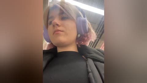 Media: A video of a young woman with fair skin, short pink hair, and large purple headphones, wearing a black top, taken from a low angle.