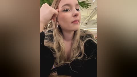 Media: Video of a fair-skinned woman with long, wavy blonde hair, wearing black clothing and dark eyeliner, with a potted plant in the background.