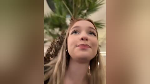 Media: Video of a young, fair-skinned woman with long blonde hair, wearing a brown dress, looking up in a room with a green palm tree in the background.