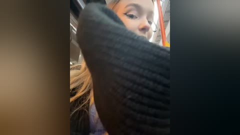 Media: Video of a young woman with light skin, long blonde hair, and light makeup, wearing a black sweater, holding a black object, possibly a smartphone, in a dimly lit, indoor setting.